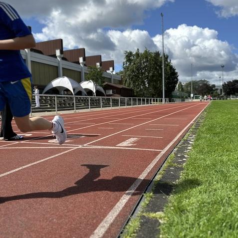 L&auml;ufer in blauer Sportkleidung auf roter Tartanbahn mit gr&uuml;nem Grasrand und bew&ouml;lktem Himmel im Hintergrund.