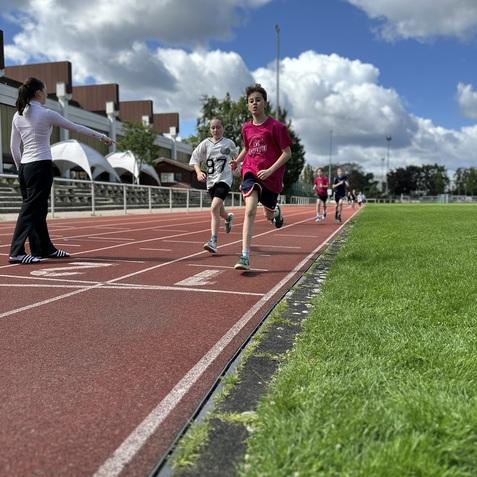 Sportlerinnen und Sportler trainieren auf einer roten Laufbahn unter einem bew&ouml;lkten Himmel.