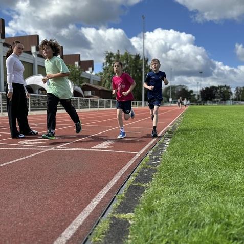Vier Kinder laufen auf einer roten Laufbahn, w&auml;hrend eine Trainerin sie beobachtet. Wolkiger Himmel im Hintergrund.