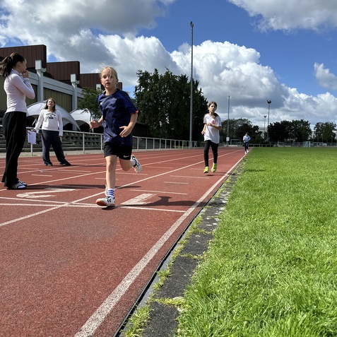Ein Junge l&auml;uft auf der Laufbahn, w&auml;hrend weitere Kinder und Erwachsene im Hintergrund stehen und beobachten.