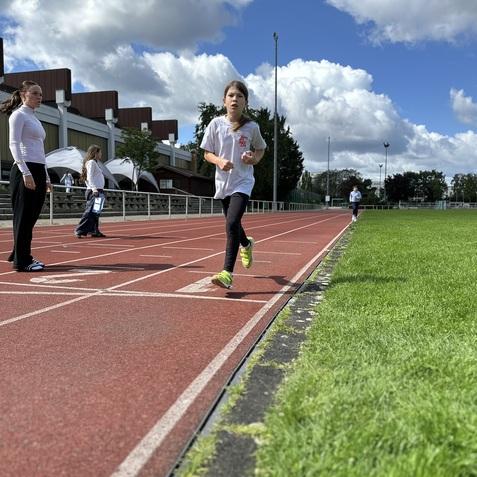 Ein M&auml;dchen l&auml;uft auf einer roten Laufbahn, w&auml;hrend Erwachsene im Hintergrund stehen. Blauer Himmel mit Wolken.