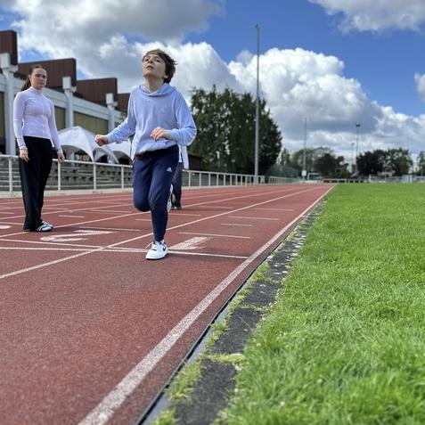 L&auml;ufer in einem hellblauen Sweatshirt sprintet auf einer roten Laufbahn, w&auml;hrend eine Trainerin daneben beobachtet.