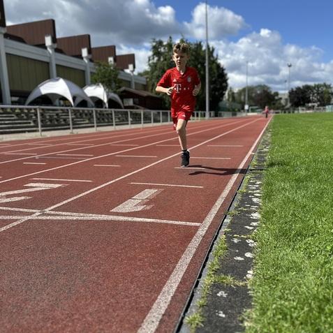 Ein Junge in rotem Sportanzug l&auml;uft auf einer Laufbahn unter blauen Himmel mit wenigen Wolken.