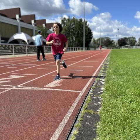 Ein Kind in einem pinken T-Shirt l&auml;uft auf einer roten Laufbahn, umgeben von Rasen und Wolken am blauen Himmel.