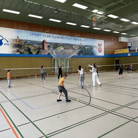 Zehn Kinder und Jugendliche spielen Badminton in einer Sporthalle mit einem gro&szlig;en Wandbanner im Hintergrund.