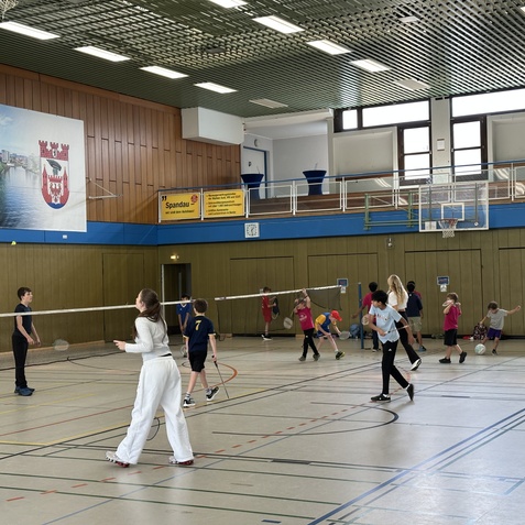 Kinder und Jugendliche spielen Badminton in einer Sporthalle mit einer Wandtafel und einem Basketballkorb.