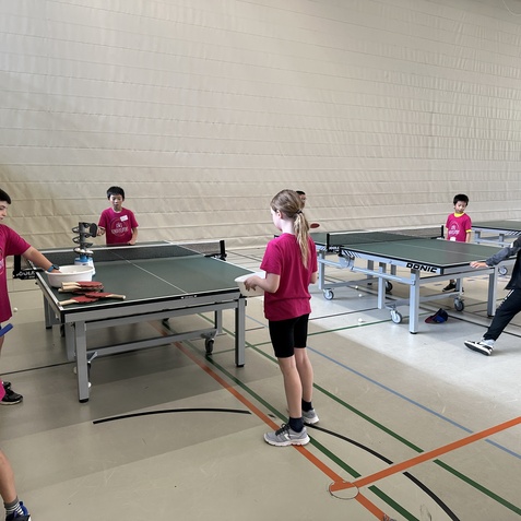 Gruppe von Kindern in pinken T-Shirts spielt Tischtennis an mehreren Tischen in einer Sporthalle.