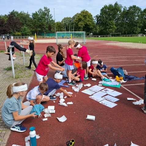 Gruppe von Kindern auf einem Sportplatz, die mit Materialien f&uuml;r ein Projekt arbeiten, einige tragen Kopfbedeckungen.