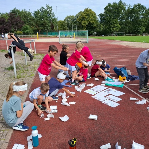 Gruppe von Kindern auf einem Sportplatz, die kreativ mit Papier und Materialien arbeiten, einige tragen Kopfbedeckungen.