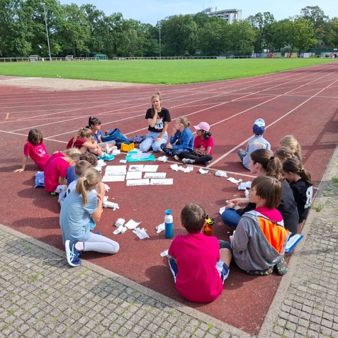 Gruppe von Kindern sitzt auf einer Laufbahn und bearbeitet Arbeitsbl&auml;tter auf dem Boden, w&auml;hrend ein Erwachsener anleitet.