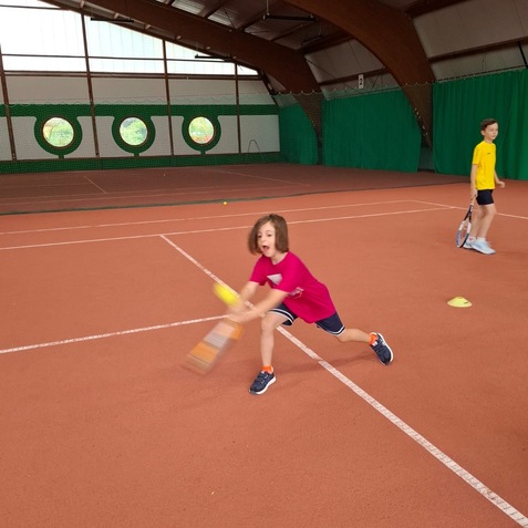 Kind in pinkem Shirt schl&auml;gt mit einem Schl&auml;ger einen Ball auf einem Tennisplatz in einer Halle. Ein weiterer Junge steht daneben.