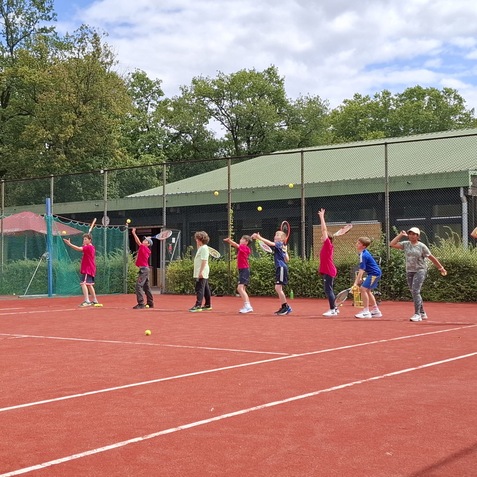 Gruppe von Kindern mit Tennisschl&auml;gern auf einem roten Tennisplatz, die B&auml;lle beim Training schlagen.