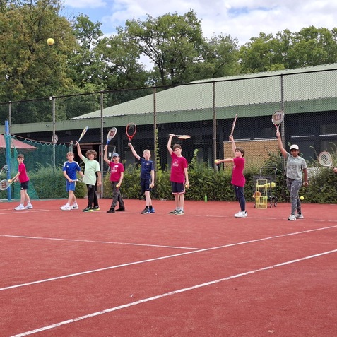 Eine Gruppe von Kindern in Sportkleidung &uuml;bt mit Tennisschl&auml;gern auf einem Tennisplatz unter blauem Himmel.