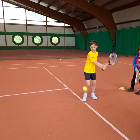 Ein Junge in einem gelben Shirt schl&auml;gt einen Tennisball auf einem Tennisplatz in einer Halle.