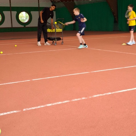 Tennistraining in Halle: Ein Trainer beobachtet einen Jungen, der einen Tennisball schlägt, während ein weiterer Junge bereitsteht.