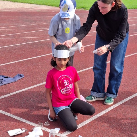 Drei Kinder auf einer Laufbahn, eines sitzt mit einer Papierbandage auf dem Kopf, w&auml;hrend zwei andere es umgeben.
