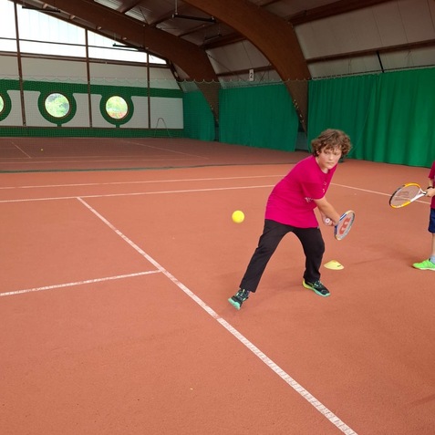 Ein Junge in rotem T-Shirt schl&auml;gt mit einem Tennisschl&auml;ger einen Ball auf einem Tennisplatz.