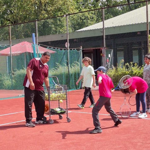 Gruppen von Kindern und einem Trainer auf einem Tennisplatz mit einem Wagen voller Tennisb&auml;lle.