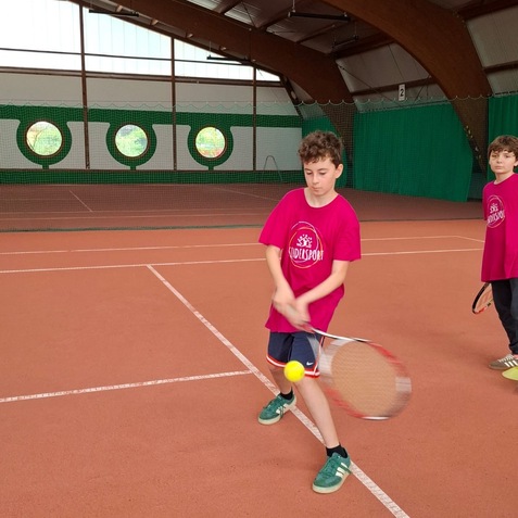 Zwei Kinder spielen Tennis in einer Halle, eines schl&auml;gt einen Ball mit einem Schl&auml;ger auf dem Sandplatz.