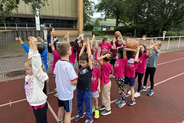 Gruppe von Kindern in pinken T-Shirts mit erhobenen H&auml;nden, einige halten Basketballs, auf einem Sportplatz.