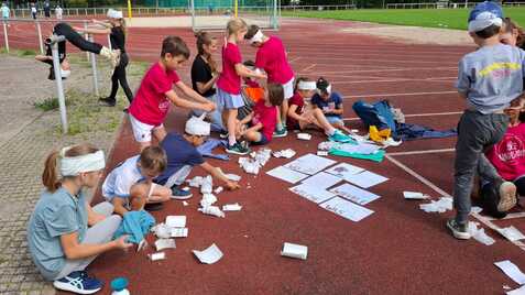 Gruppe von Kindern auf einem Sportplatz, die mit Materialien für ein Projekt arbeiten, einige tragen Kopfbedeckungen.