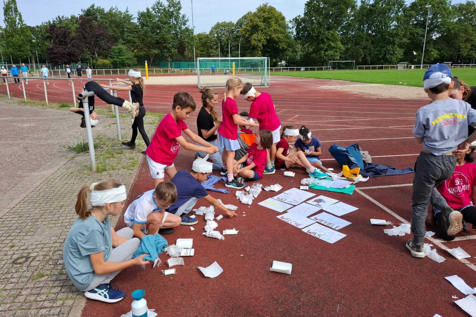 Gruppe von Kindern auf einem Sportplatz, die mit Materialien f&uuml;r ein Projekt arbeiten, einige tragen Kopfbedeckungen.