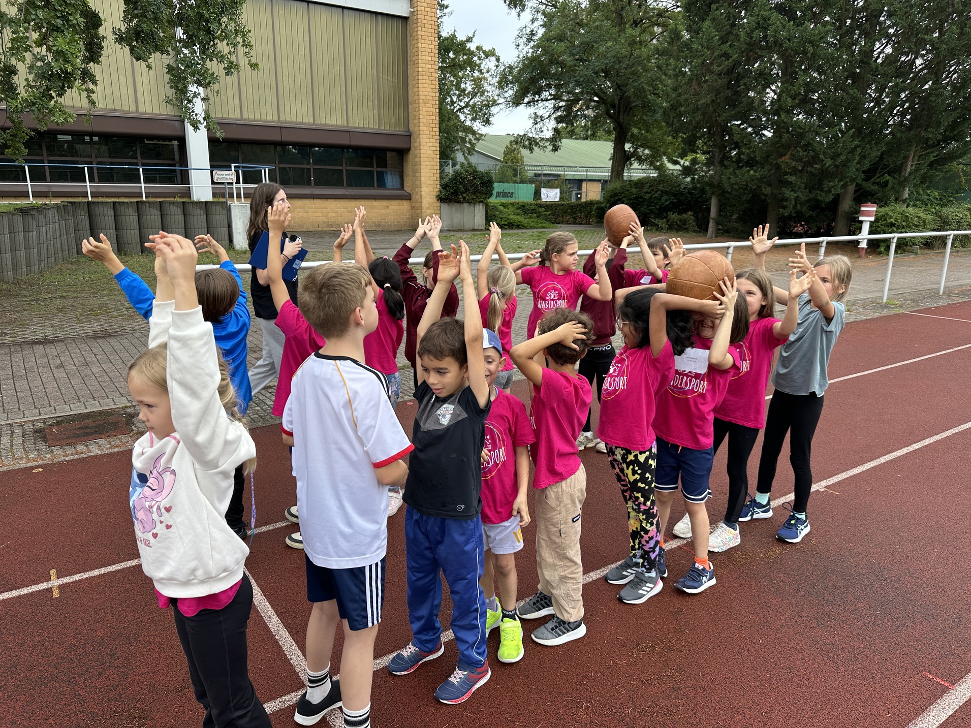 Gruppe von Kindern in pinken T-Shirts mit erhobenen Händen, einige halten Basketballs, auf einem Sportplatz.