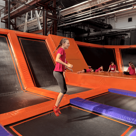 M&auml;dchen springt auf einem Trampolin in einem Park, umgeben von orangefarbenen und lilanen Trampolinen.