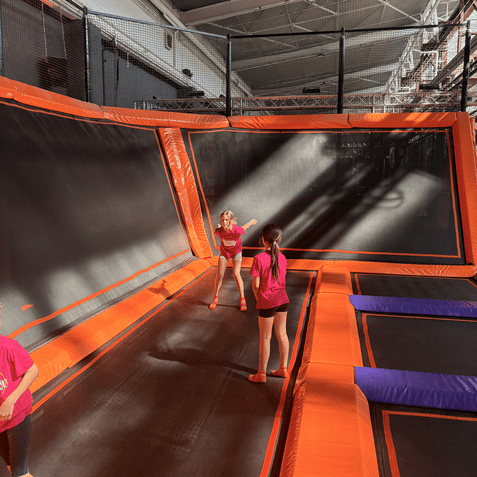 Drei Kinder in T-Shirts springen auf einer Trampolinfläche mit orangefarbenem Rahmen in einer Indoor-Trampolinhalle.