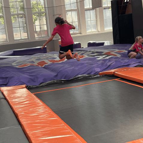 Ein Mädchen springt auf einem Trampolin, ein weiteres Mädchen sitzt daneben auf einer weichen Landefläche.