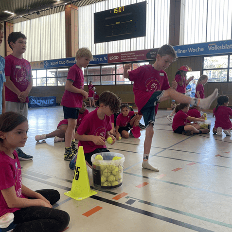 Kinder in rosa T-Shirts stehen und sitzen in einer Sporthalle, w&auml;hrend sie an einem Spiel mit Tennisb&auml;llen teilnehmen.