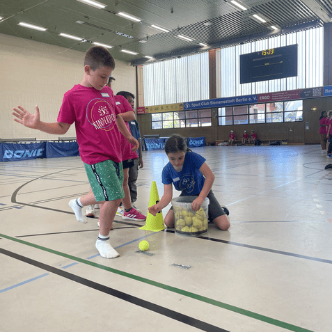Zwei Kinder stehen in einer Sporthalle; eines platziert einen Tennisball neben einem Hütchen, das andere hält einen Behälter mit Bällen.