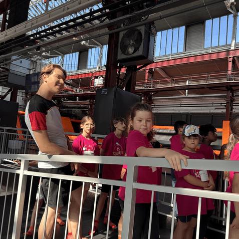 Gruppe von Kindern in pinken T-Shirts wartet an einem Geländer in einer Halle mit hohen Fenstern.