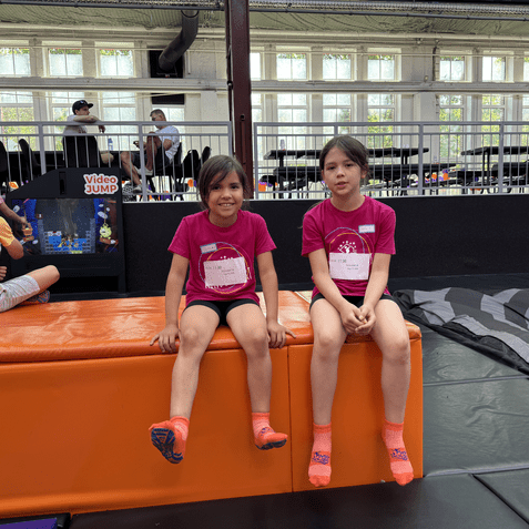 Zwei Mädchen in pinken T-Shirts sitzen auf einer orangefarbenen Matte in einer Trampolinhalle. Im Hintergrund spielen Kinder.