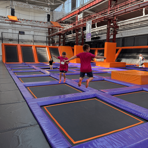 Zwei Kinder springen auf lila Trampolinen in einer gro&szlig;en, hellen Trampolinhalle mit orangefarbenen Akzenten.