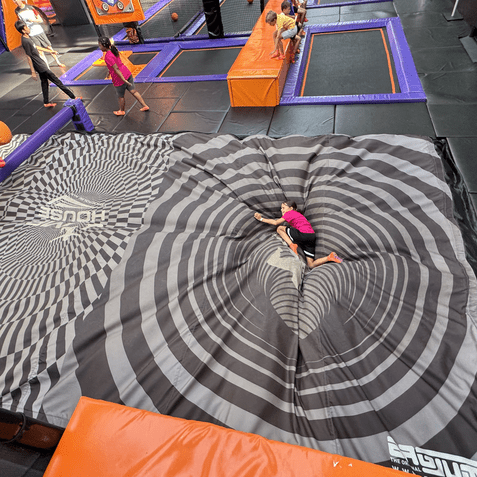 Kind in pinkem Shirt liegt auf einer spiralförmigen Matte in einem Trampolinpark, umgeben von weiteren spielenden Kindern.