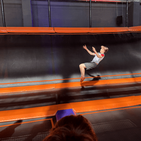 Junge springt auf einem orangefarbenen Trampolin, während ein Mädchen in rosa T-Shirt zuschaut.