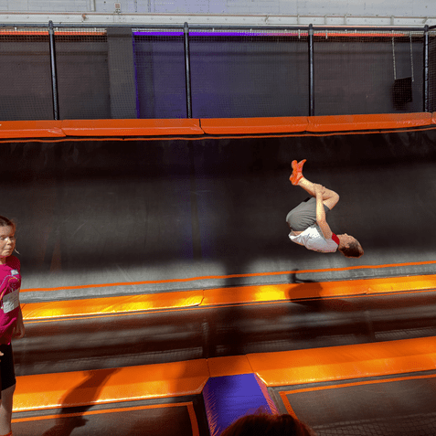 Ein Junge führt einen Rückwärtssalto auf einem Trampolin aus, während ein Mädchen daneben steht.