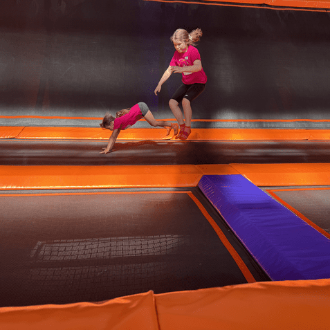 Zwei Kinder in pinken Shirts springen auf einem Trampolinpark mit orangefarbenen und lila Sprungflächen.