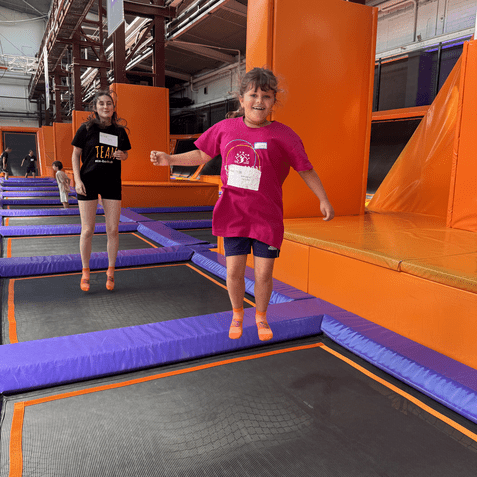 Zwei Kinder springen auf Trampolinen in einer Trampolinhalle mit orangefarbenen Wänden und lila Randpolstern.