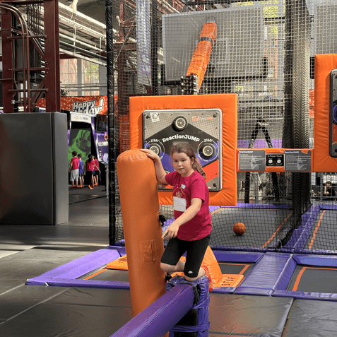 Kind in rotem T-Shirt steht auf einem blauen Balken an einer Trampolin-Anlage mit orangefarbenen Ger&auml;ten.
