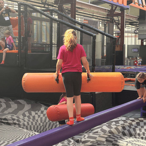 M&auml;dchen balanciert auf einer Schaumstoffbahn und h&auml;lt zwei aufblasbare Rollen in einer Trampolinhalle.