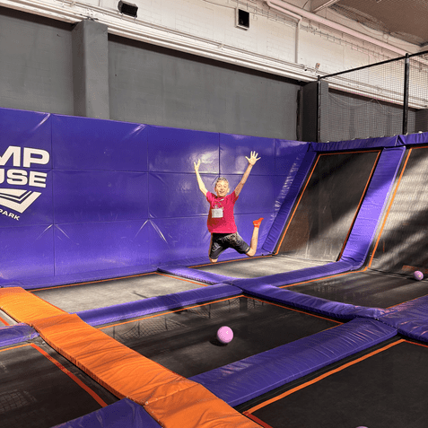 Ein Kind springt auf einer Trampolinfl&auml;che und hat die Arme in die Luft gehoben, w&auml;hrend pinke B&auml;lle im Hintergrund liegen.