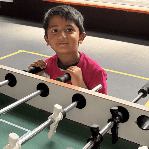 Junge mit braunen Haaren steht an einem Tischfu&szlig;ballspiel und schaut l&auml;chelnd in die Kamera.