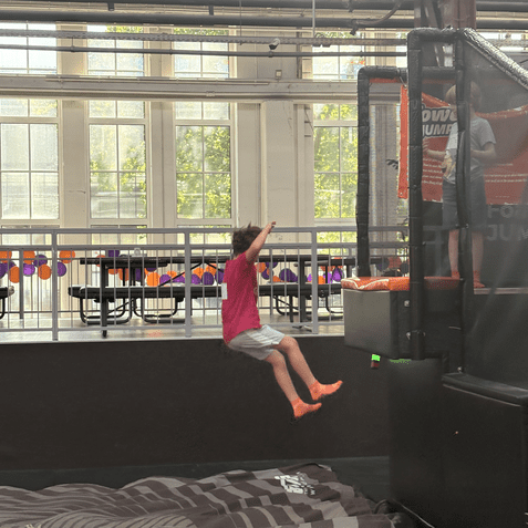 Ein Kind in rotem T-Shirt springt von einer Plattform hinunter auf eine schwarze Sprungmatte in einer Trampolinhalle.