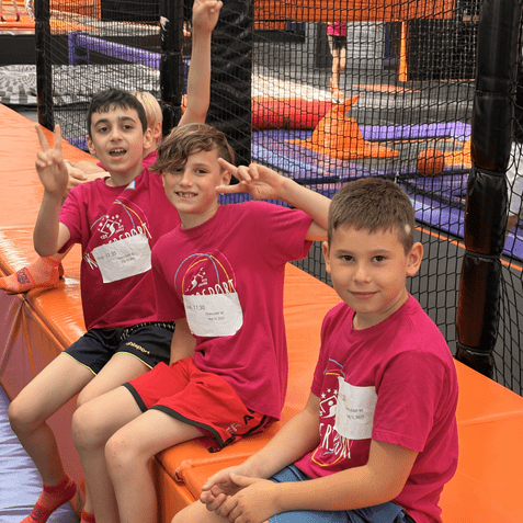 Drei Kinder mit pinken T-Shirts sitzen auf einer orangefarbenen Bank in einem Trampolinpark und zeigen das Peace-Zeichen.