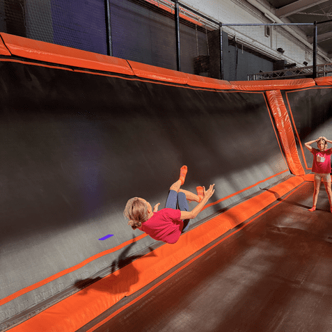 M&auml;dchen springt in eine Trampolinhalle, mit orangefarbenen Trampolinen und einer anderen Person im Hintergrund.