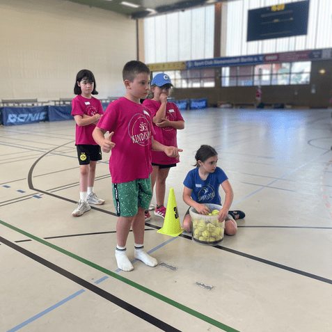 Vier Kinder in bunten Shirts stehen und sitzen in einer Sporthalle mit einem Behälter voller Bälle.