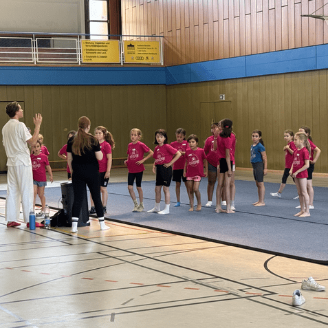Gruppe von Kindern in pinken T-Shirts hört einer Trainerin in einer Turnhalle aufmerksam zu.