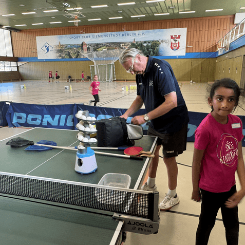 Ein Trainer sortiert Ausrüstung neben einem Tischtennistisch, während Kinder in pinken Shirts trainieren.
